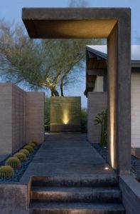 Geometric fixture arching over pathway with light shining against the surface of this fixture. At the end of the pathway is a light shining up against a coal cement wall.